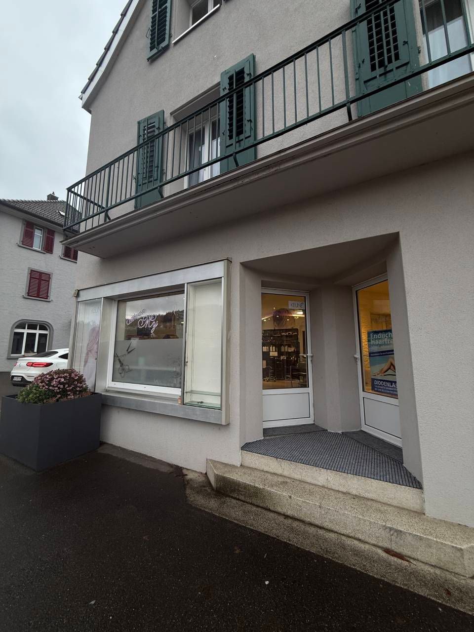 Aussenansicht eines Gebäudes mit einem Ladenlokal. Graue Fassade, Balkon, Schaufenster und eine Tür, aus der Licht scheint.