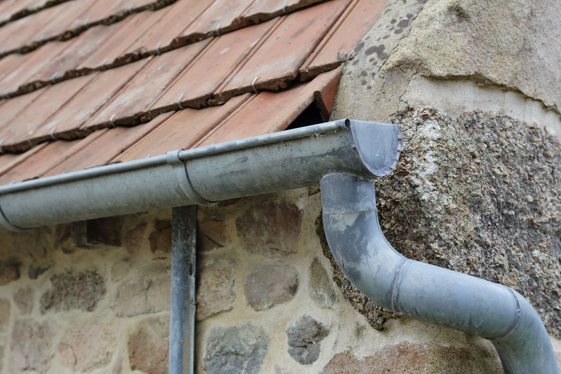 Gouttières et descentes pluviales grises sur un bâtiment en pierre avec un toit de tuiles rouges.