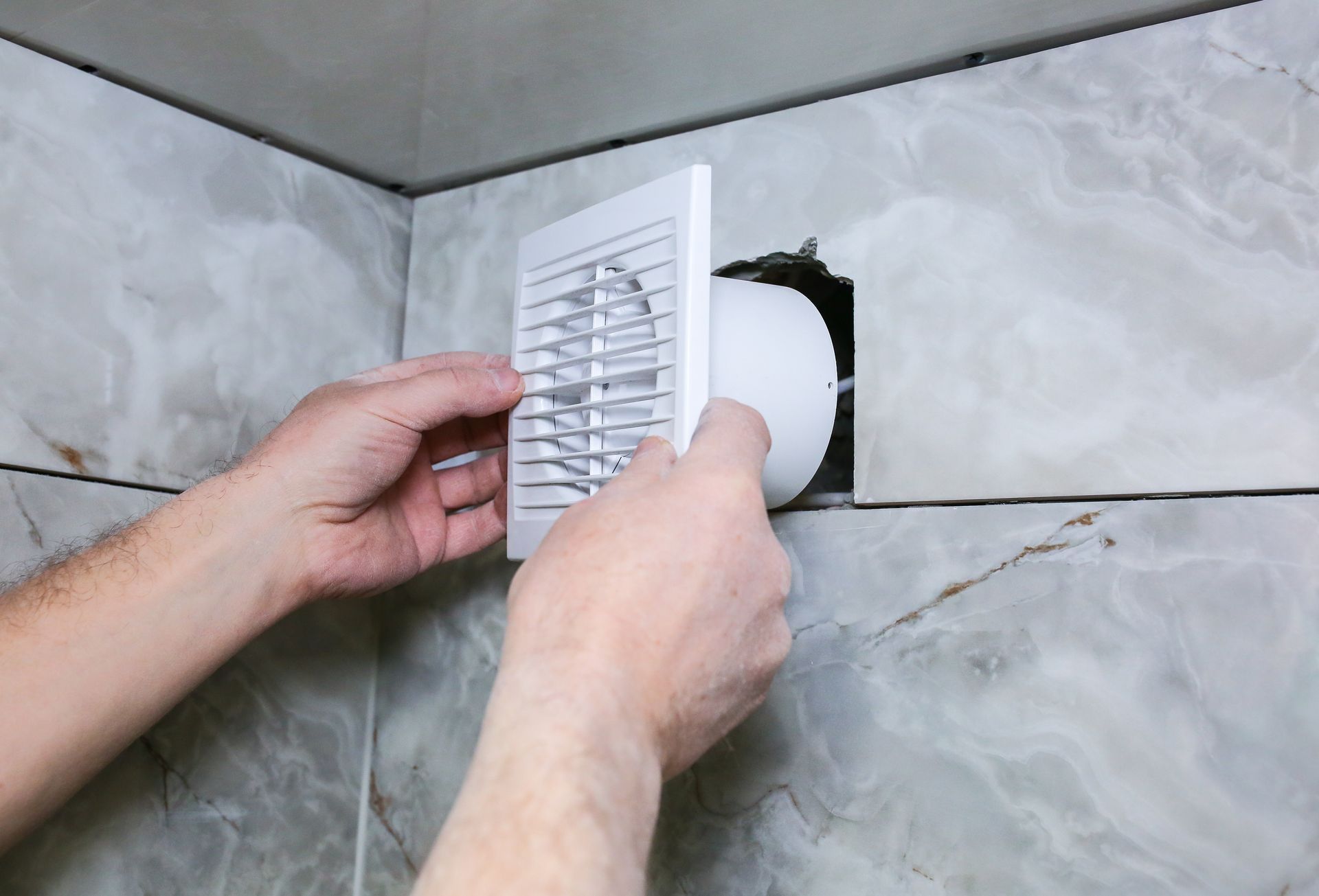 Des mains installent une grille d'aération blanche sur un mur de salle de bain carrelé de carreaux imitation marbre.