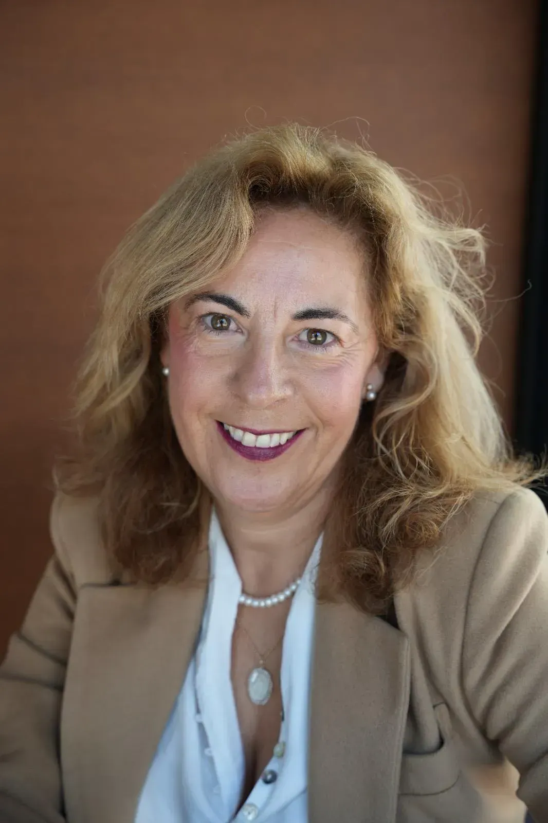 Una mujer con un blazer color canela sonríe, llevando un collar de perlas, en un interior con un fondo marrón.