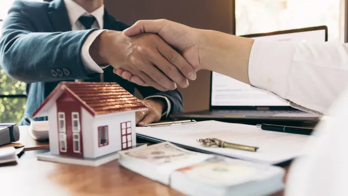 Dos personas se dan la mano sobre una casa en miniatura, dinero y documentos sobre un escritorio.