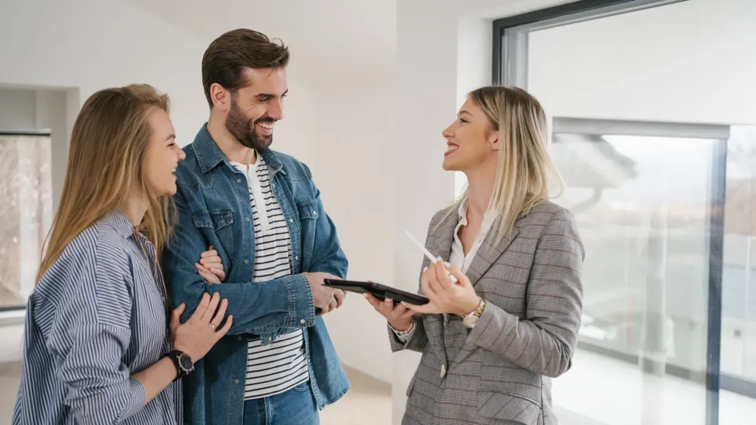 Agente inmobiliario muestra documentación a una pareja sonriente en una casa moderna y luminosa.