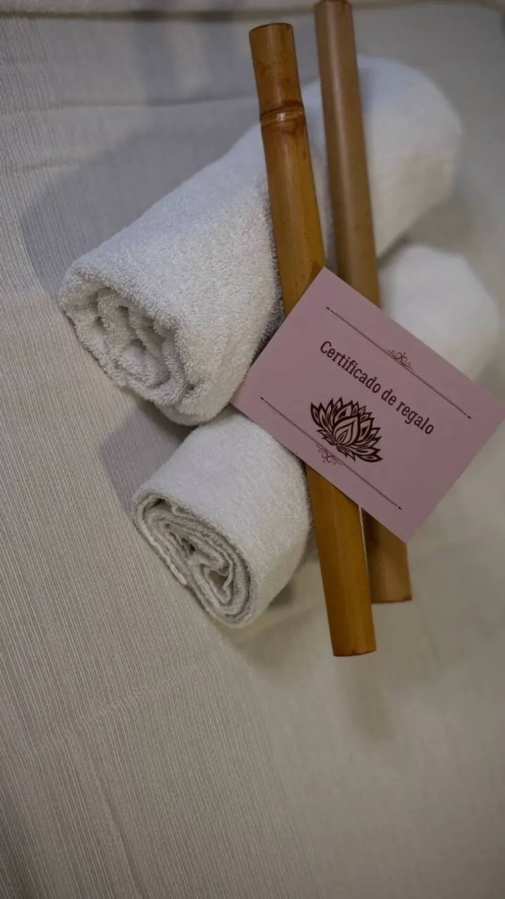 Dos toallas blancas enrolladas, acompañadas de palitos de bambú y un certificado de regalo rosa sobre una superficie blanca.