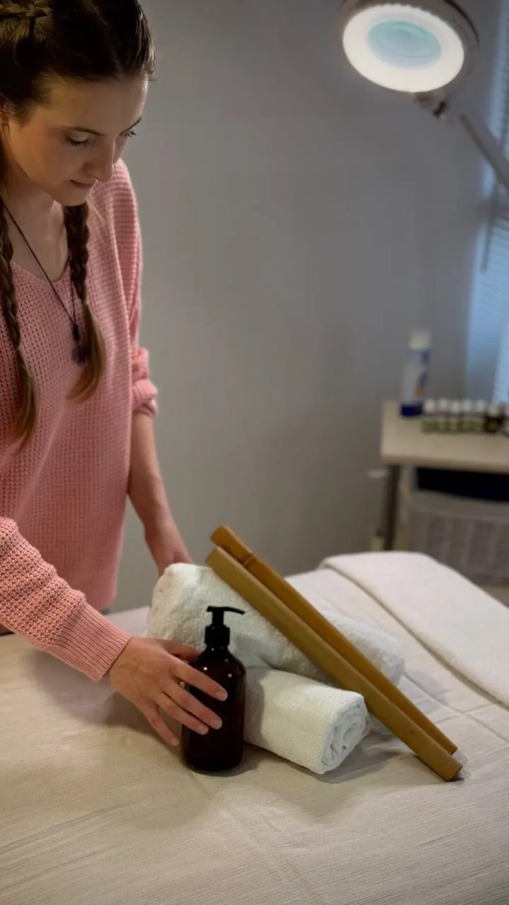 Una persona con un suéter rosa coloca toallas y un frasco con dosificador sobre una camilla de masaje