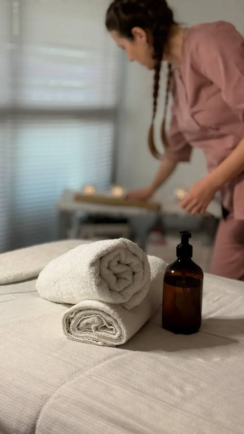 Una persona con uniforme rosa prepara una sala de spa con toallas enrolladas y aceite sobre una camilla de masaje.
