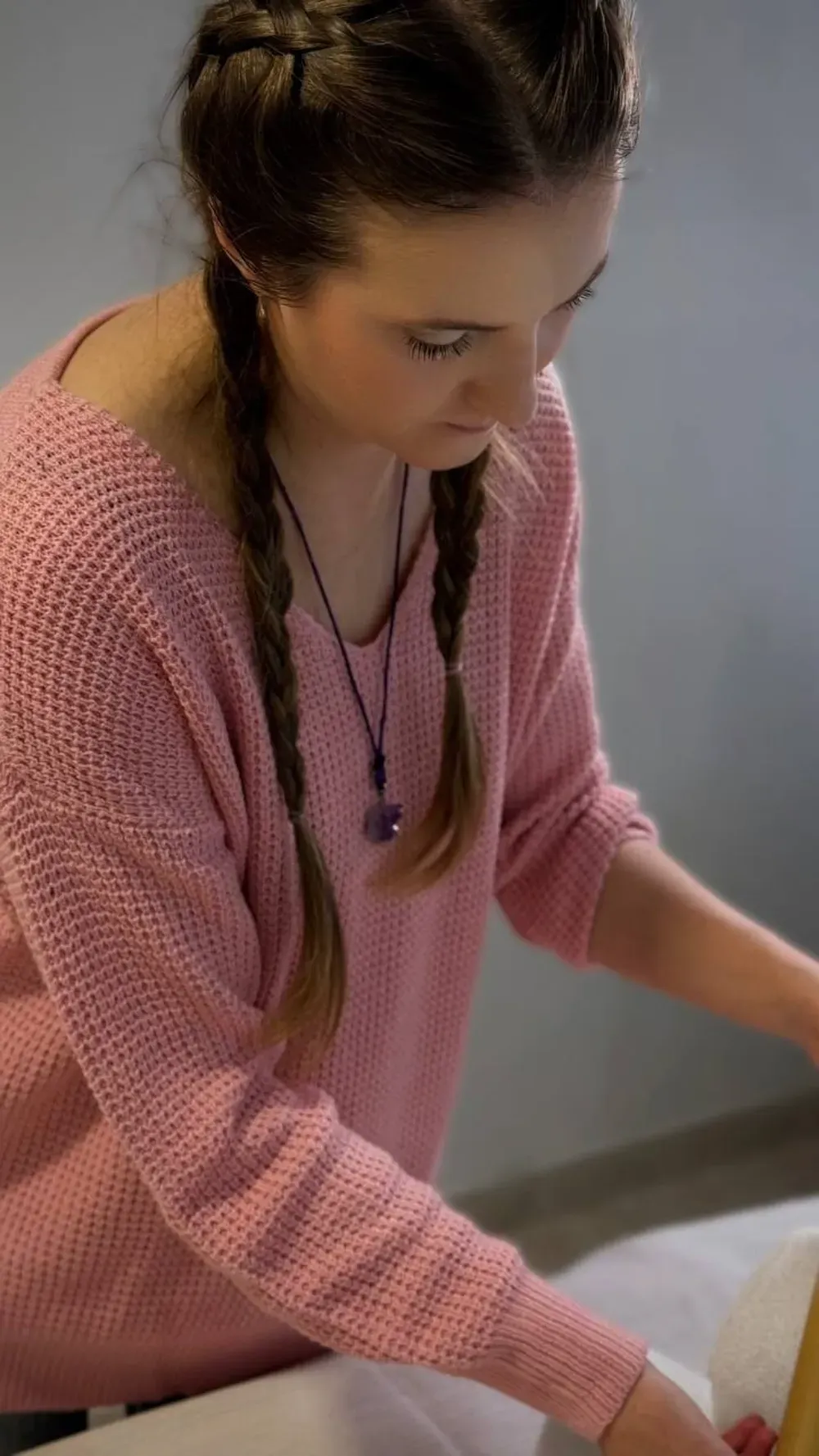 Una persona con el pelo trenzado, que lleva un jersey de punto rosa y un collar con colgante, se inclina hacia adelante