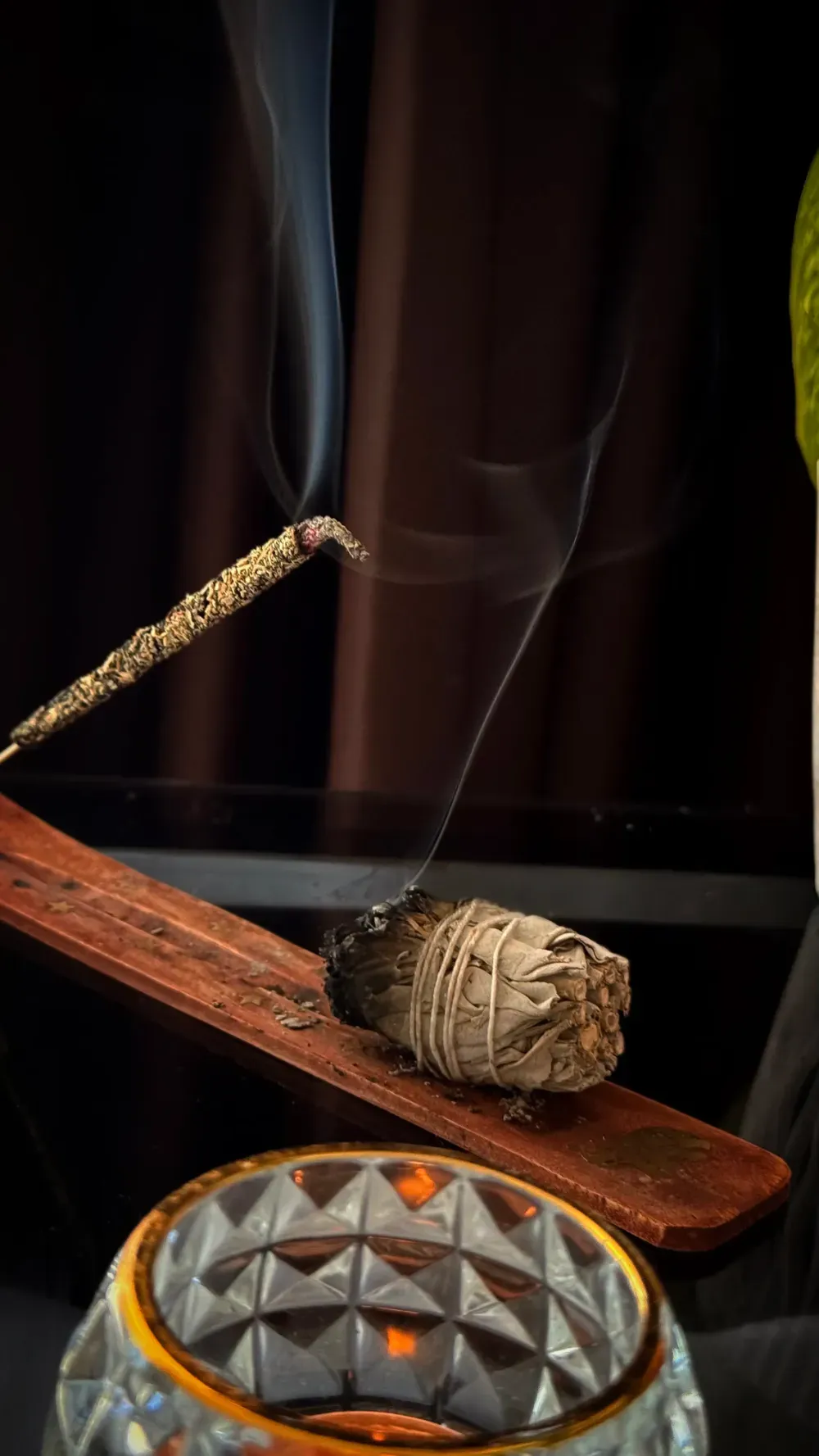 Un manojo de salvia humeante reposa sobre un soporte de madera junto a una varita de incienso encendida