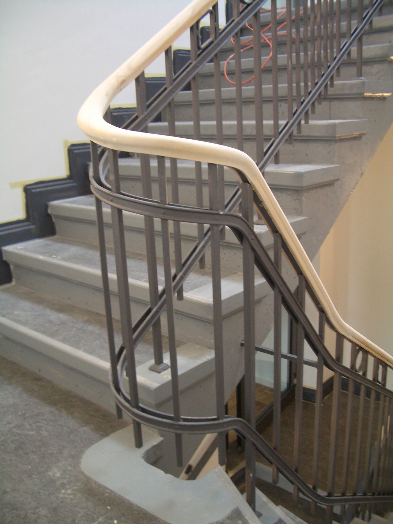 Treppe mit grauen Stufen, schwarzem Metallgeländer und hellem Holzhandlauf.