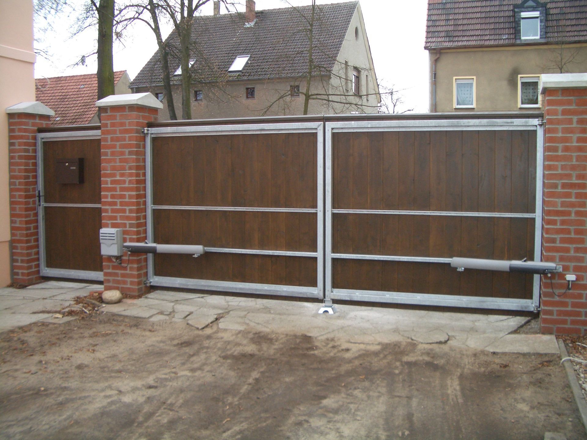 Braunes, hölzernes Doppeltor mit Metallverzierungen, Backsteinsäulen und einer Auffahrt.