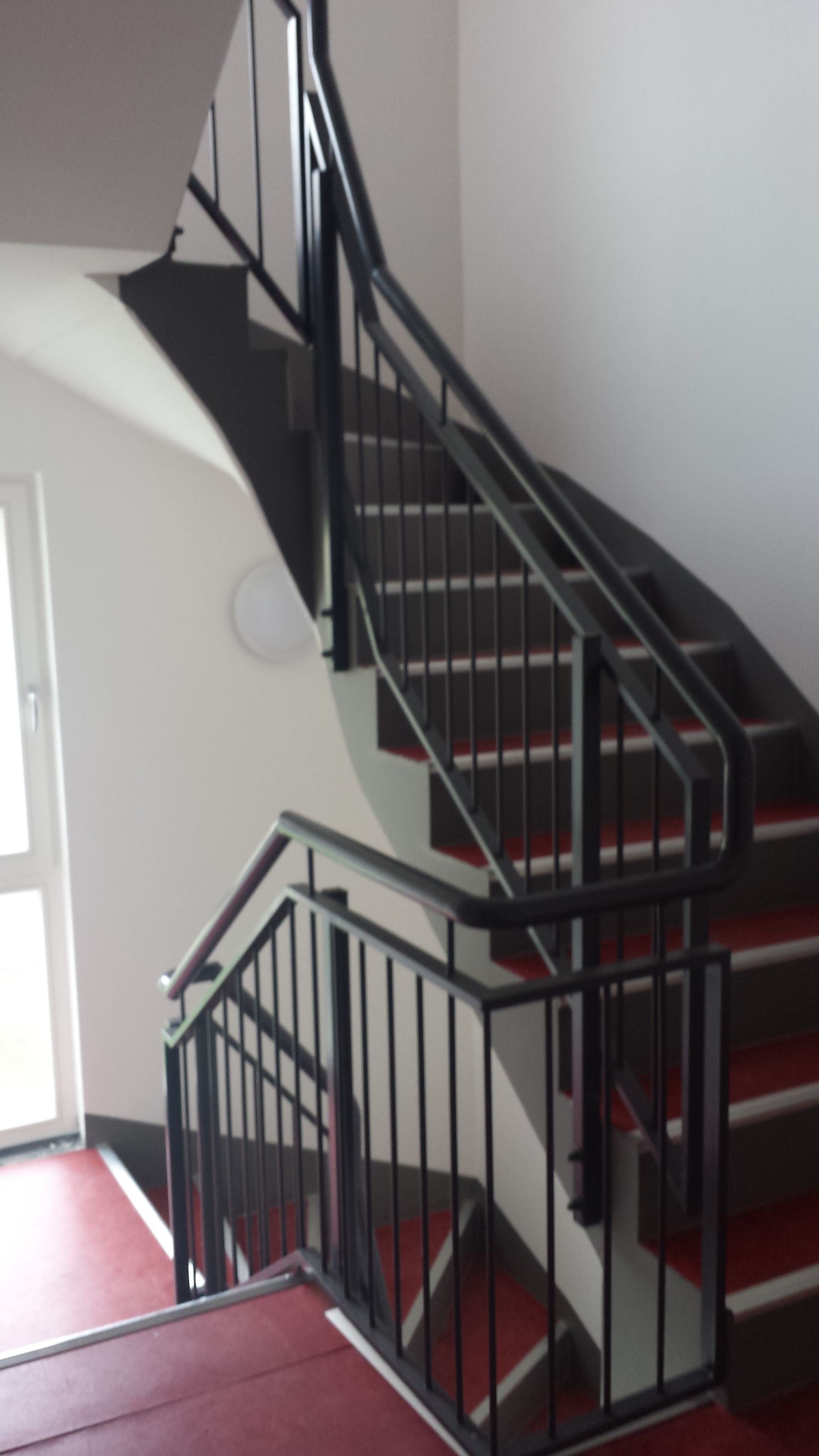 Treppe mit schwarzem Metallgeländer und roten Stufen vor weißen Wänden.