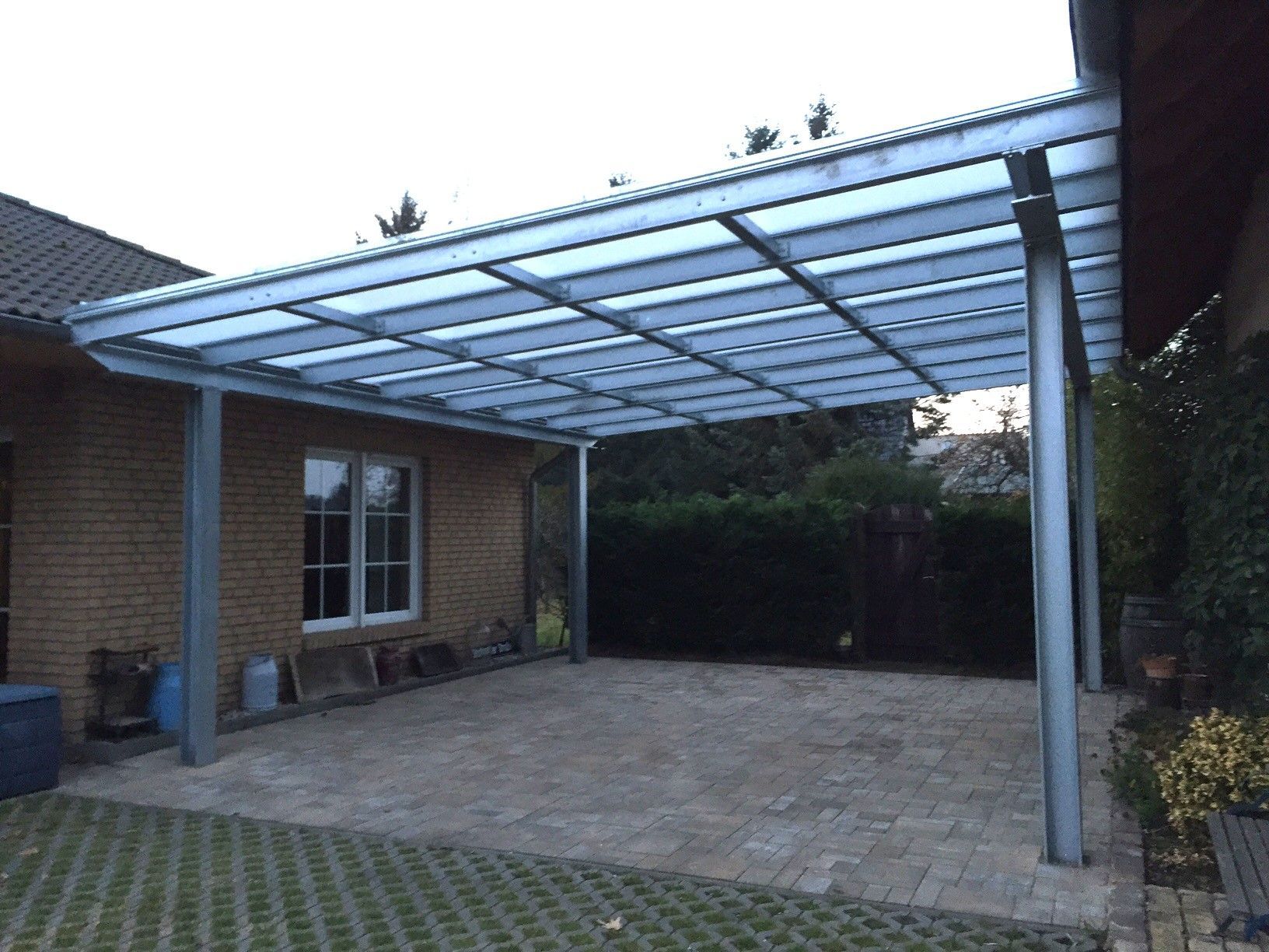 Carport mit Metallrahmen, transparentem Dach und Betonboden neben einem Backsteingebäude.