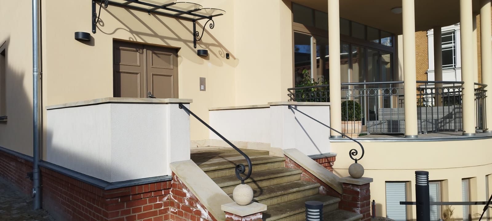 Außenansicht eines Gebäudes mit Treppe. Braune Tür. Beige Wände. Schwarzes Geländer mit Zierelementen.