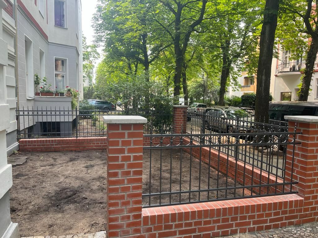 Ein Zaun aus Ziegelsteinen und Schmiedeeisen umschließt ein unbefestigtes Stück Land vor einem Gebäude mit Bäumen.