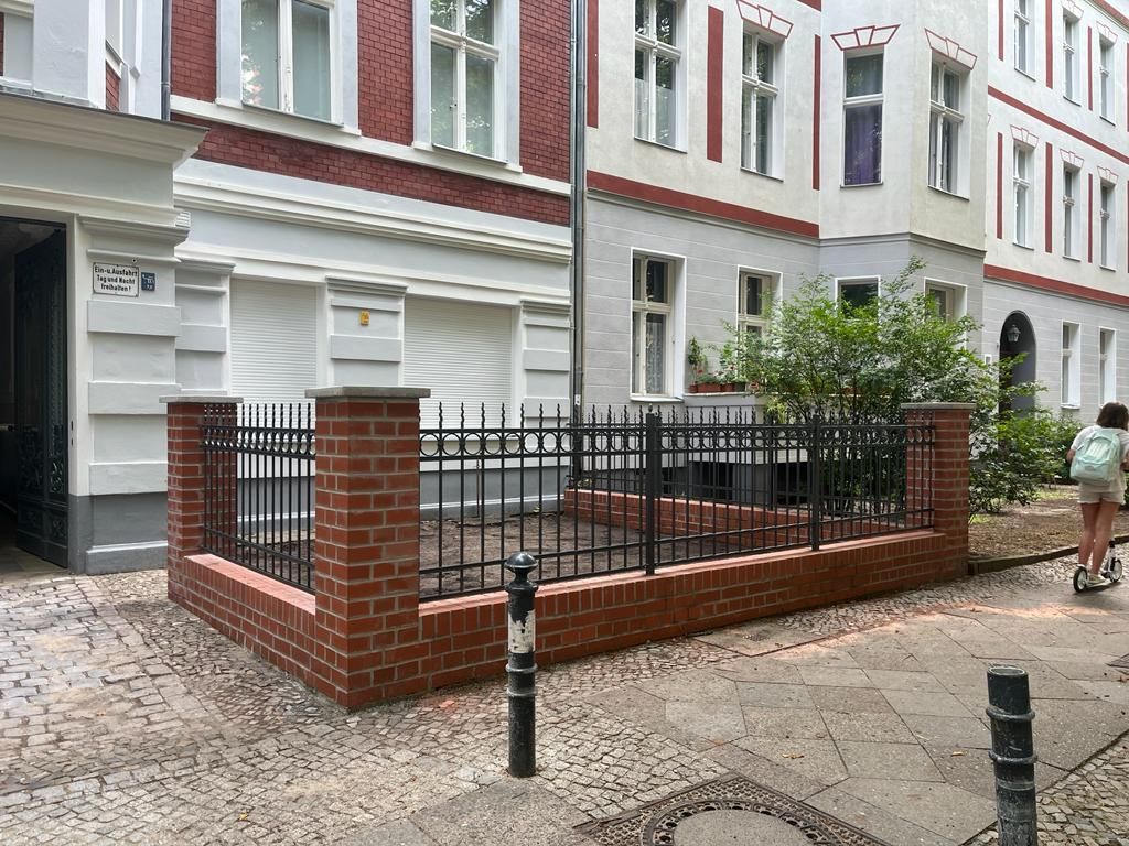 Ein gemauerter Zaun mit schwarzem Metallzaun vor einem Gebäude.
