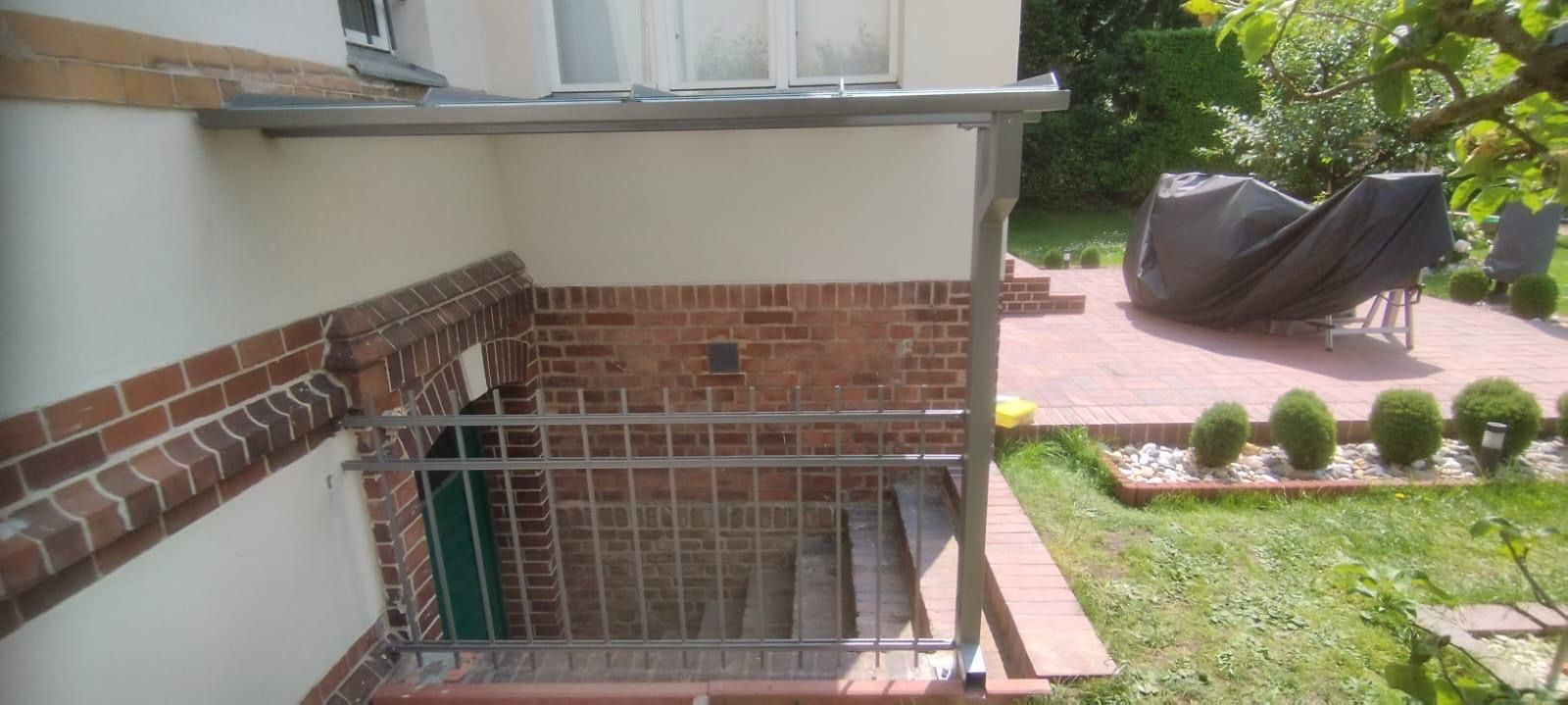 Eingang mit Treppe, Backsteinmauer und Metallgeländer. Eine Terrasse mit überdachten Gartenmöbeln ist sichtbar.