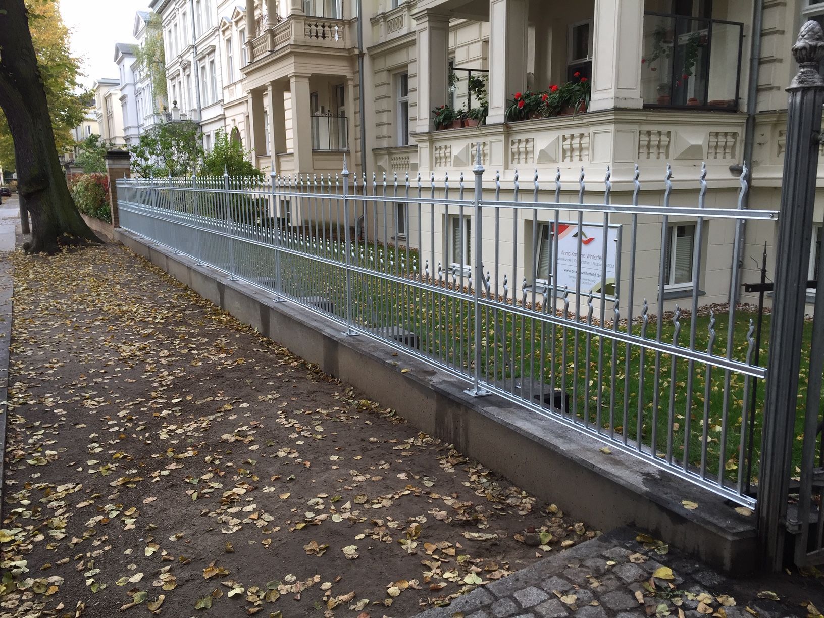 Ein Metallzaun entlang eines Gehwegs trennt die Straße von einem Gebäude mit Fenstern und Balkonen.