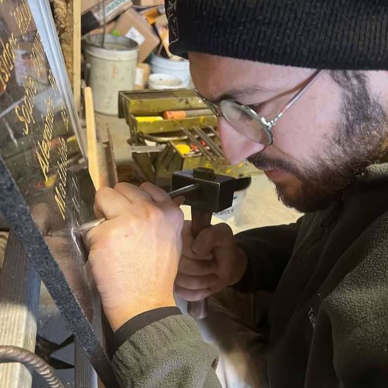 Une personne portant des lunettes et un bonnet noir travaille avec soin sur un petit objet, assise à un établi avec des outils à proximité.