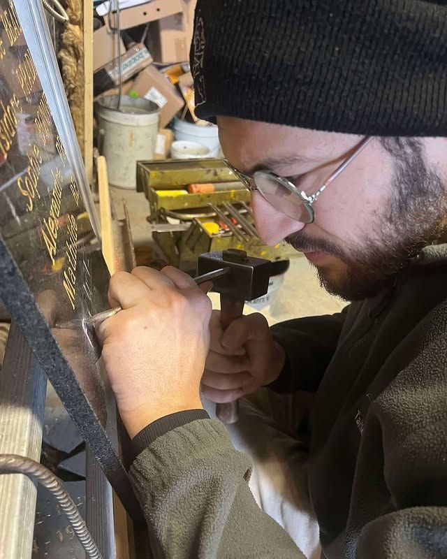 Un homme portant un bonnet noir et des lunettes travaille avec des outils à un établi d'atelier.