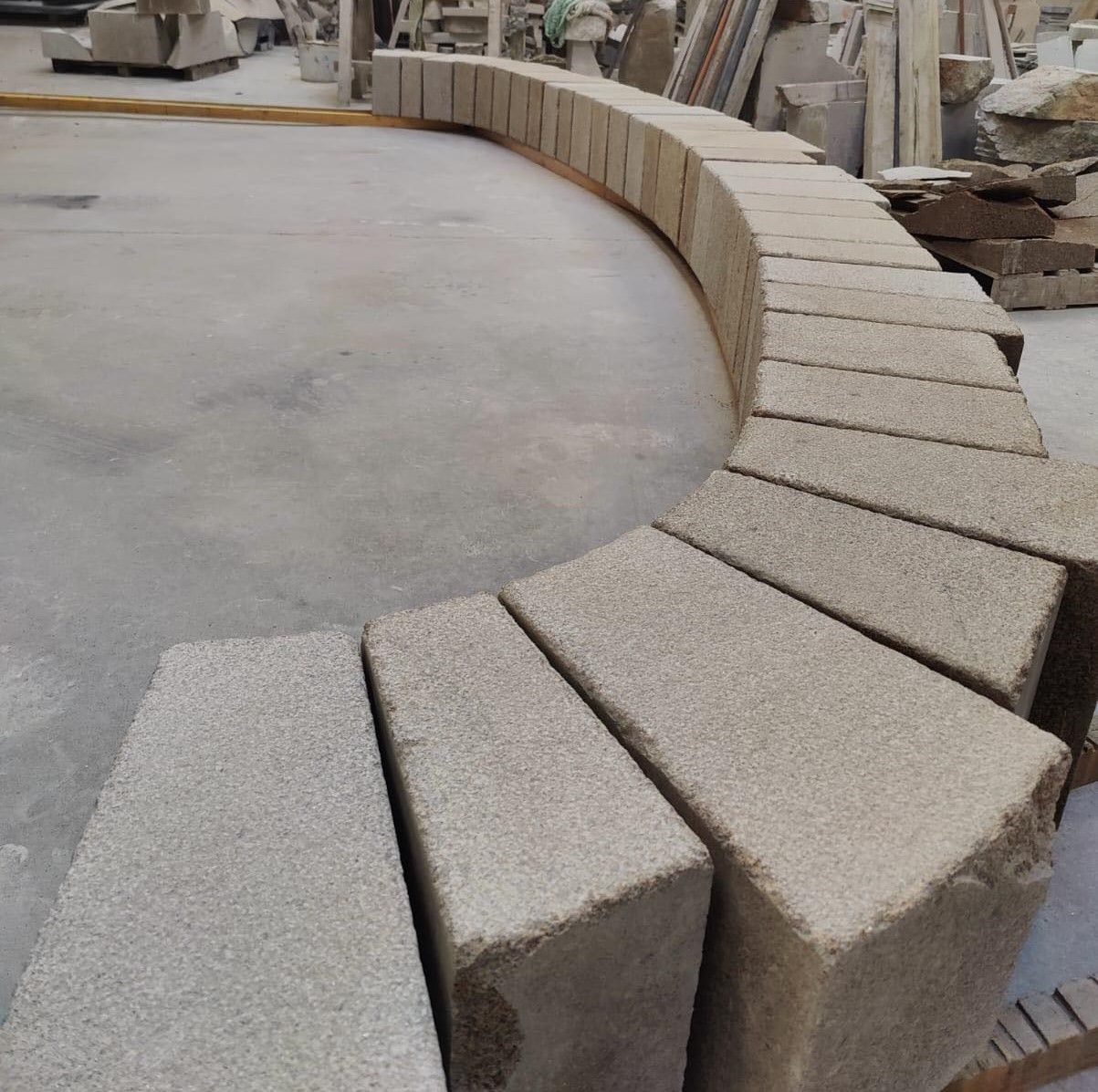 Un banc en blocs de pierre incurvés est en cours de fabrication dans un atelier, avec des blocs gris et beiges disposés en arc de cercle.