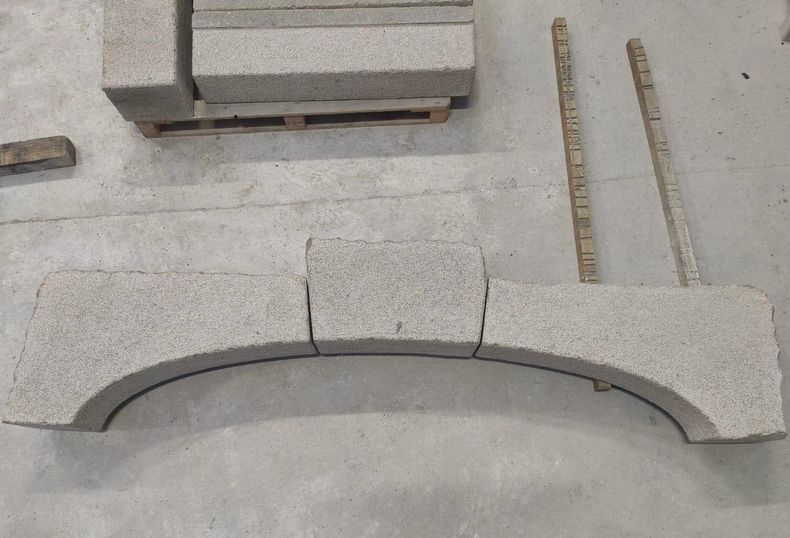 Bloc d'arche en pierre grise avec une découpe courbe, présenté parmi d'autres pièces de pierre dans un atelier ou sur un chantier.