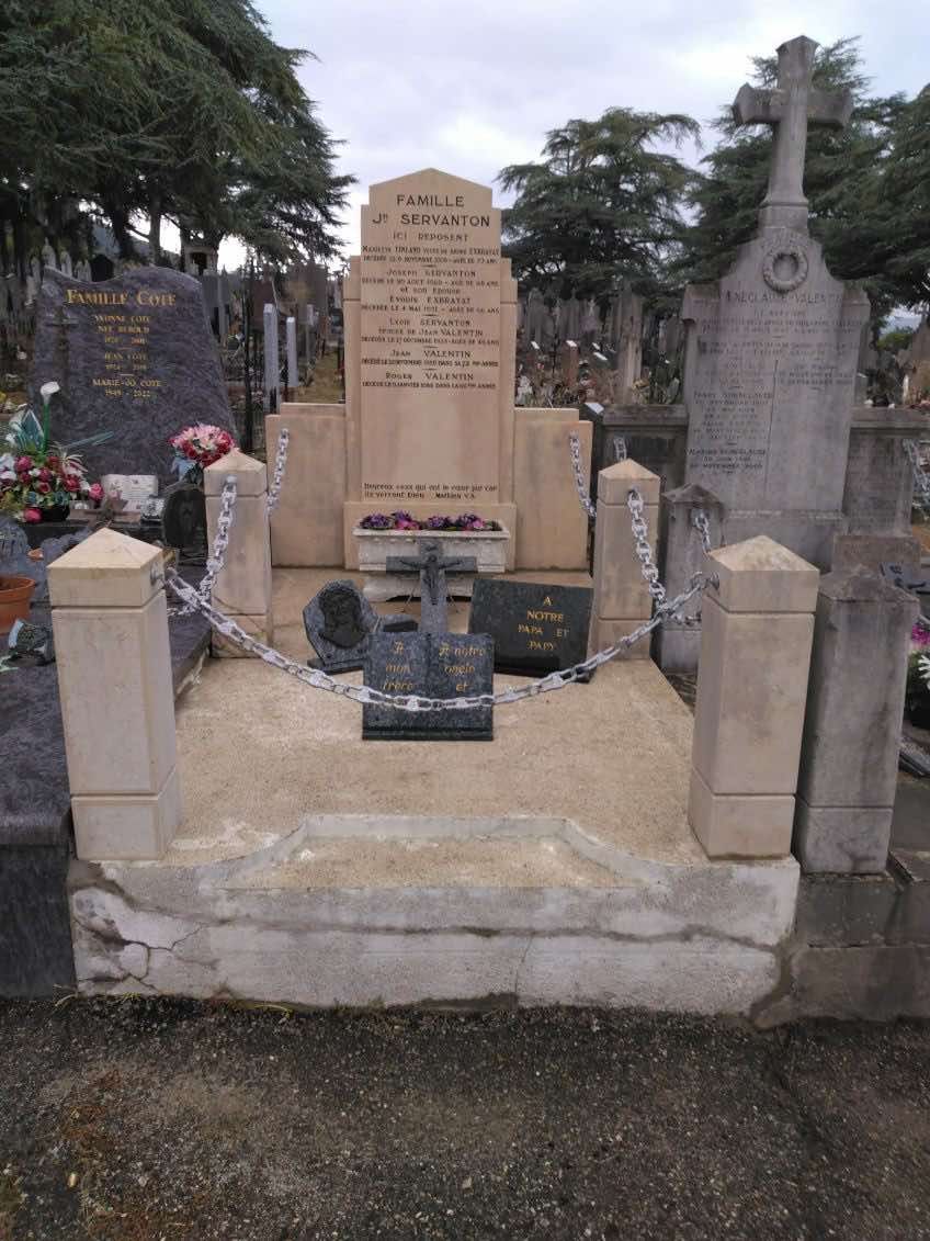 Monument funéraire composé de pierres tombales beiges et grises dans un cimetière, entouré de fleurs et de poteaux en pierre.