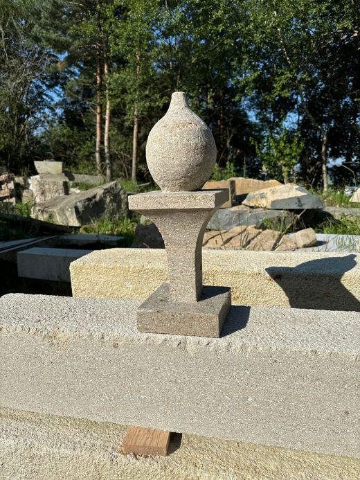 Sculpture en forme d'urne de pierre sur un piédestal dans un jardin extérieur ensoleillé, agrémenté de rochers et d'arbres.