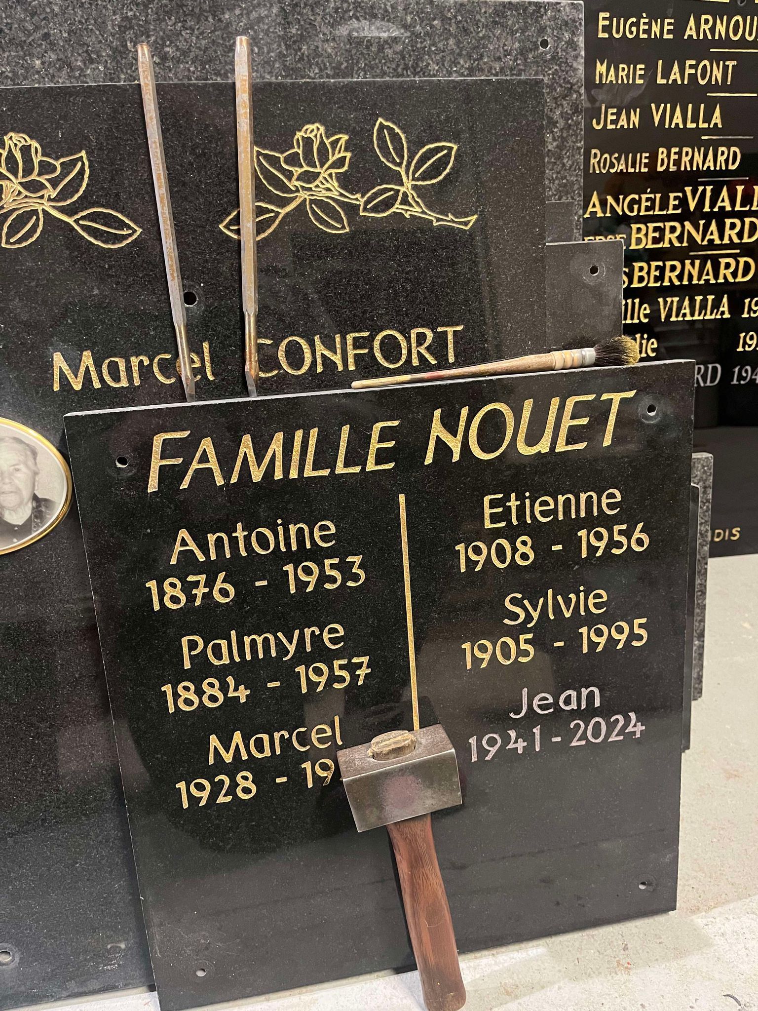 Pierre tombale familiale en granit noir, ornée de lettres dorées et de petites gravures florales, surmontée d'une croix en bois.