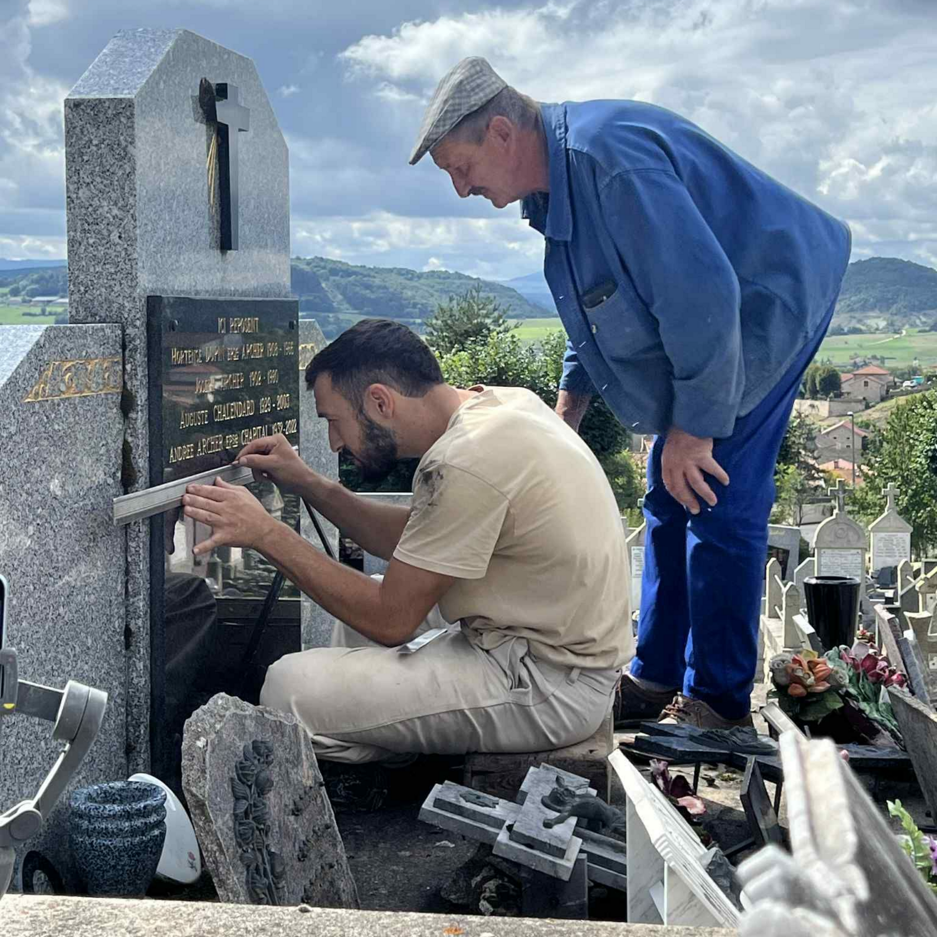 Deux hommes réparent une pierre tombale dans un cimetière, l'un agenouillé et l'autre debout, avec un village à flanc de colline en arrière-plan.