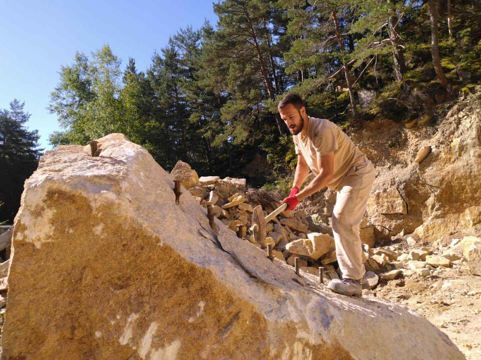 Un homme utilise une masse pour casser un gros rocher à l'extérieur, sur un chantier de construction rocailleux.