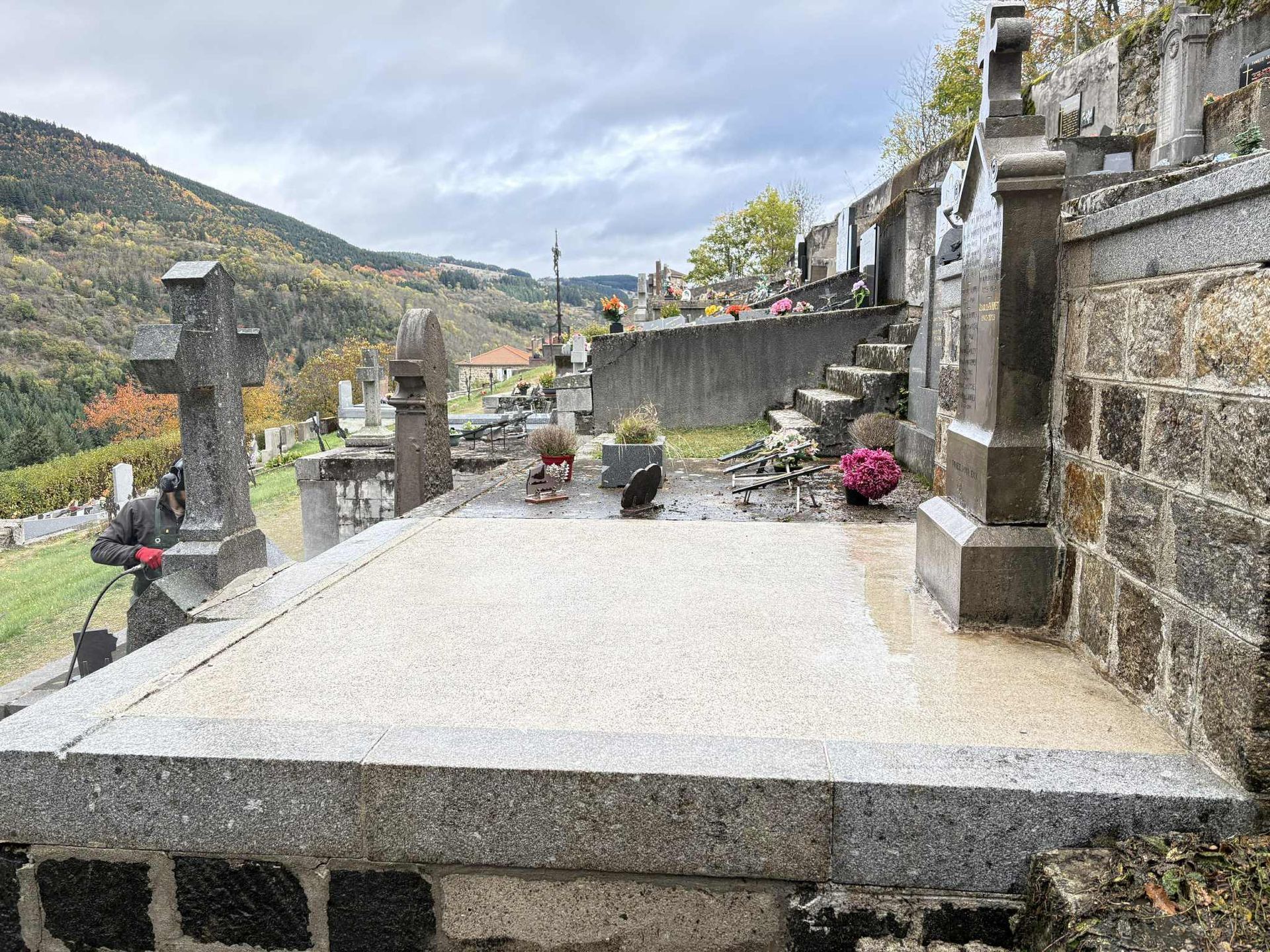 Allée en terrasse de pierre dans un cimetière à flanc de colline, agrémentée de plusieurs compositions florales et offrant une vue sur les montagnes.