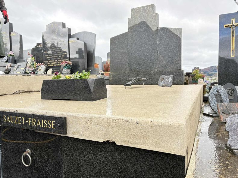 Pierre tombale dans un cimetière, avec un tombeau beige et une stèle en granit foncé parmi d'autres monuments.