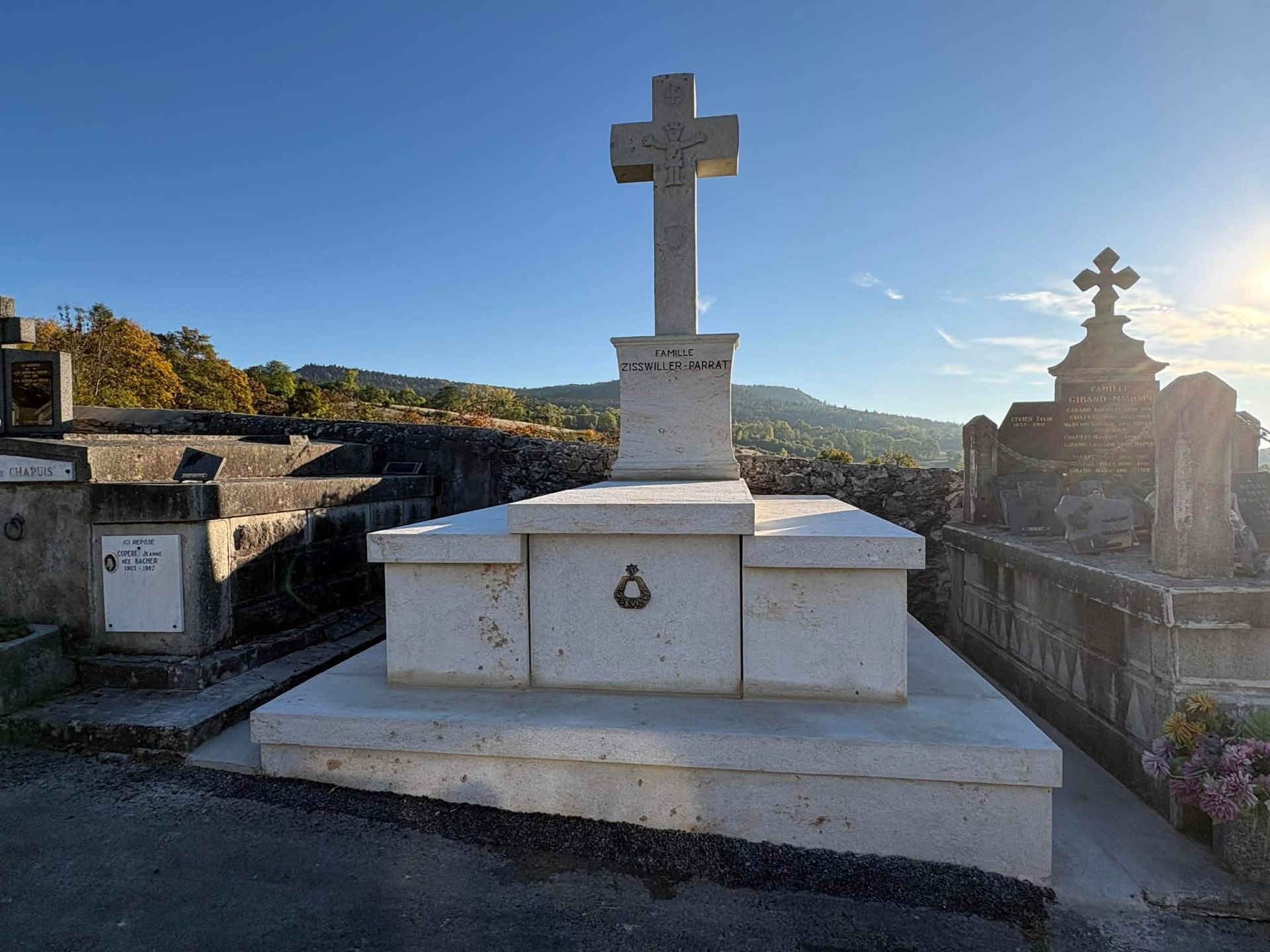 Croix en pierre blanche dans un cimetière, avec des montagnes et un ciel bleu en arrière-plan