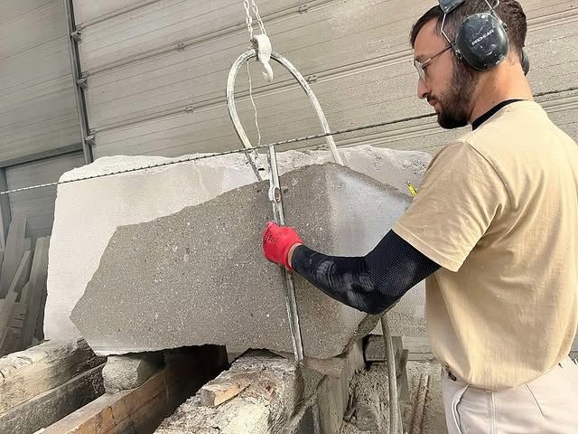 Un ouvrier portant des protections auditives et des gants découpe un gros bloc de pierre dans un atelier.