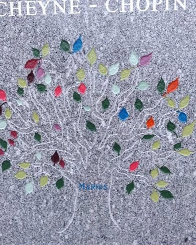 Gravure sur pierre tombale d'un arbre aux branches blanches et aux feuilles multicolores sur fond gris.