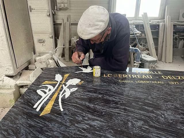 Ouvrier peignant une pierre commémorative noire dans un atelier, avec des lettres dorées et un motif jaune et blanc