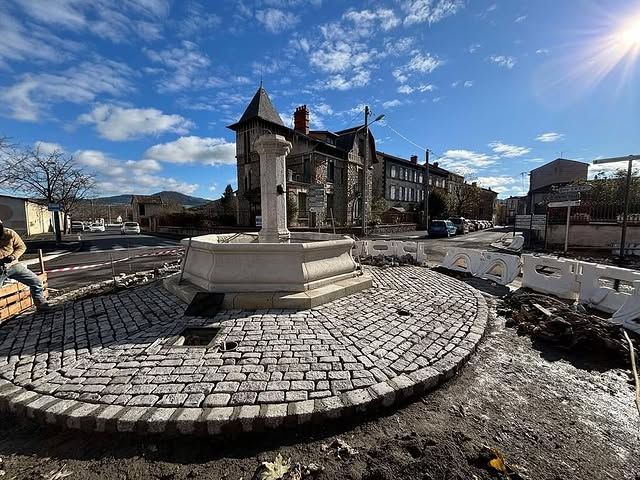 Fontaine en pierre sur une place pavée, à côté d'un bâtiment historique, sous un ciel bleu.