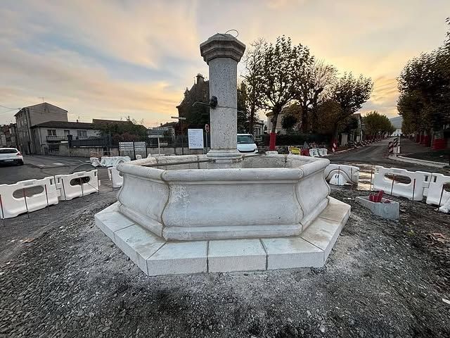 Base de fontaine en pierre vide dans un chantier au coucher du soleil, avec des barrières et des arbres en arrière-plan.