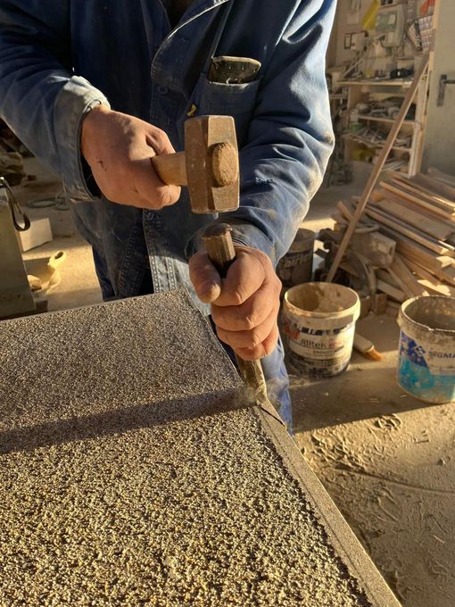 Un ouvrier taille un bloc de pierre sur un chantier, avec des outils et du gravier à proximité.