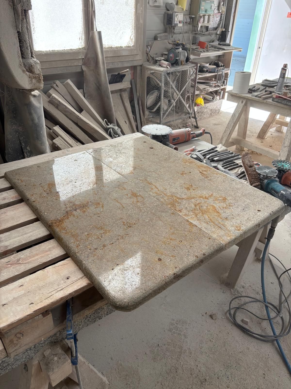 Dalle de pierre sur un établi dans un atelier poussiéreux, avec des outils, des planches de bois et une chaise à proximité.
