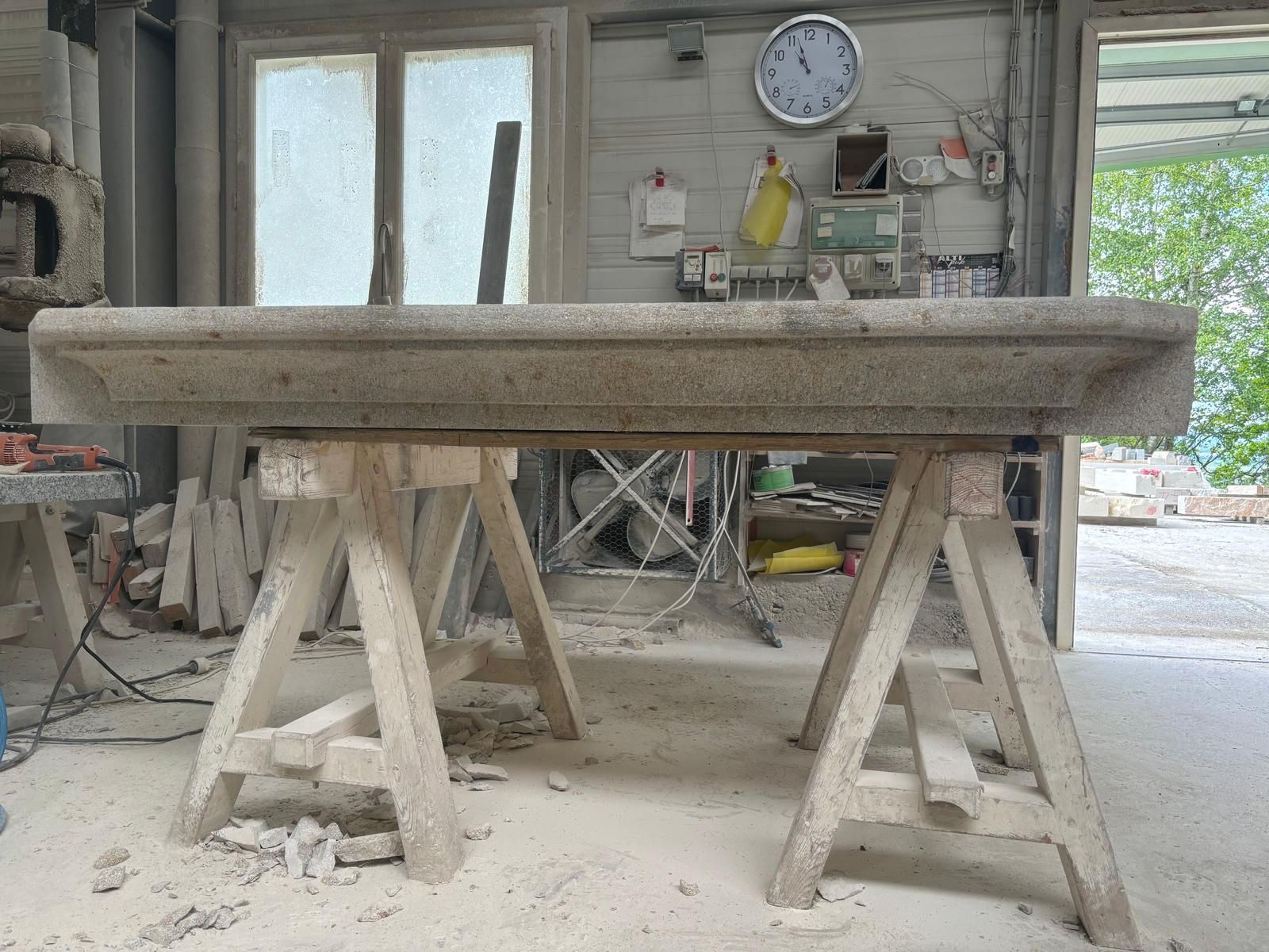 Dalle de pierre posée sur des tréteaux dans un atelier poussiéreux, avec des outils et une horloge au mur.