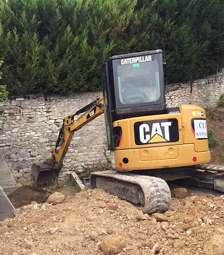 Une pelleteuse Caterpillar jaune creuse devant un mur de pierre et des buissons verts.