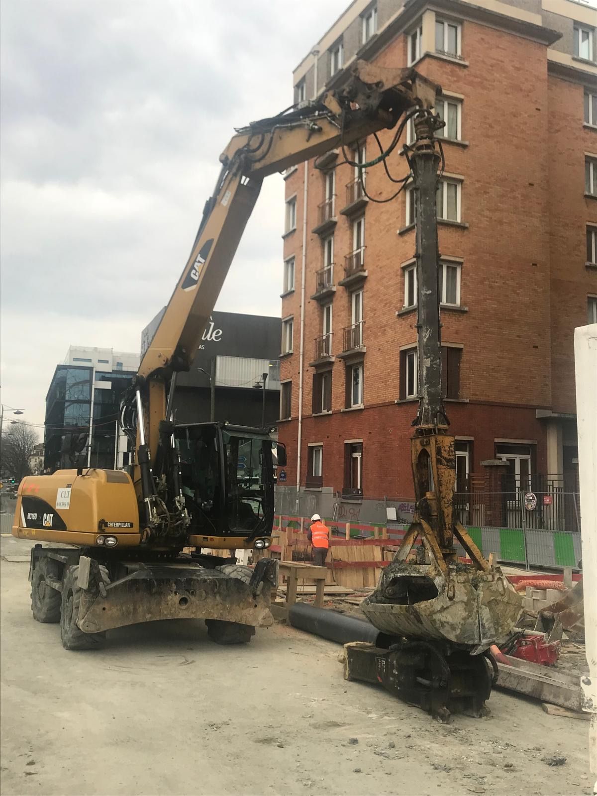 Une excavatrice jaune équipée d'un dispositif de forage est à l'œuvre sur un chantier de construction près d'un bâtiment en briques.