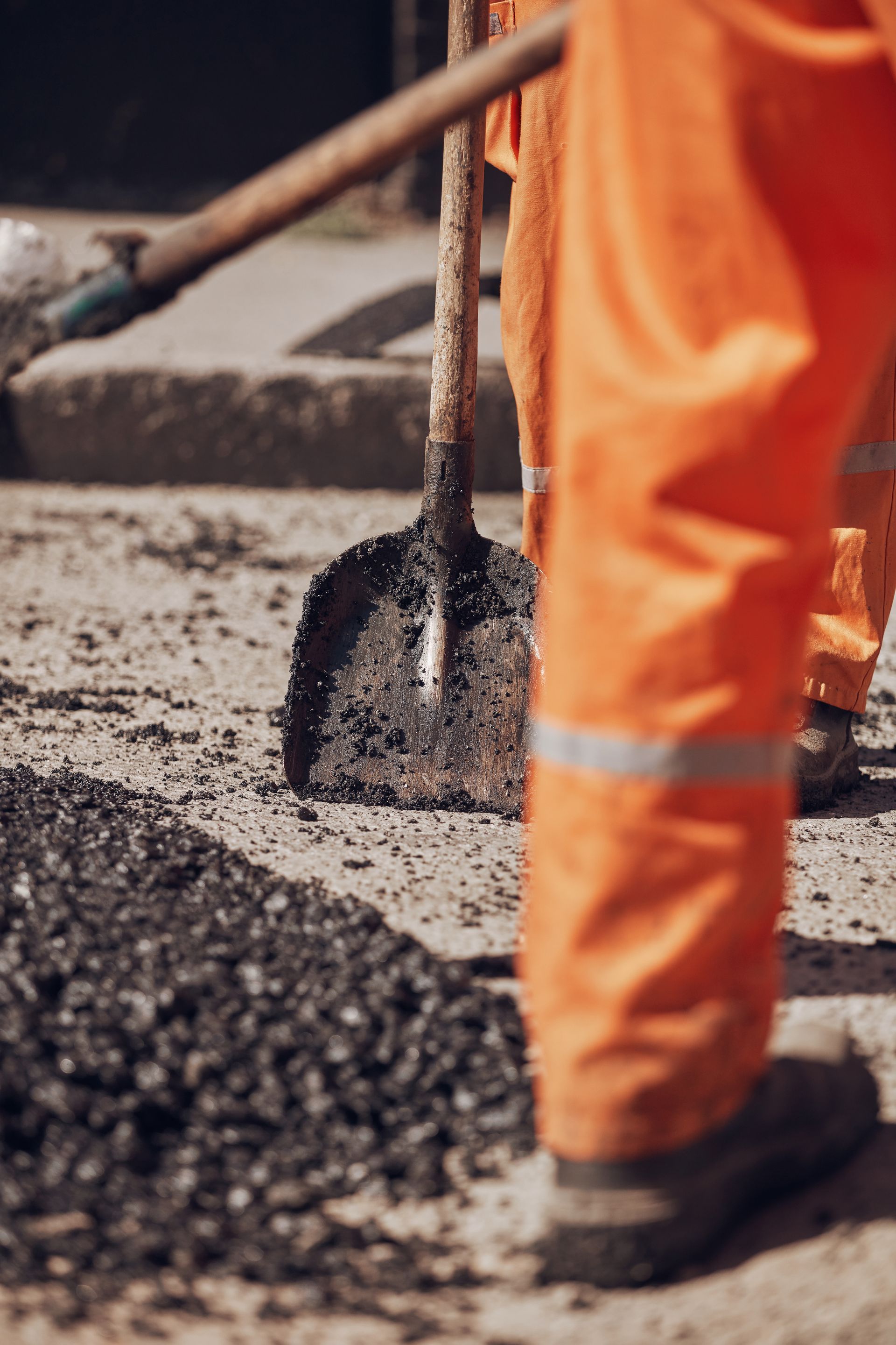 Une personne en pantalon de travail orange utilise une pelle pour étaler de l'asphalte sur une route.