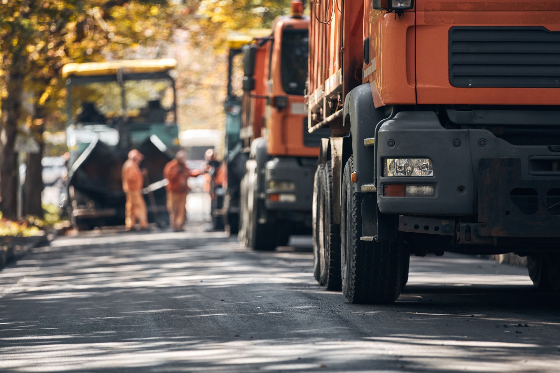 Scène de travaux de pavage : des camions orange bordent la route ; des ouvriers en gilets orange se trouvent près de la machine à paver.
