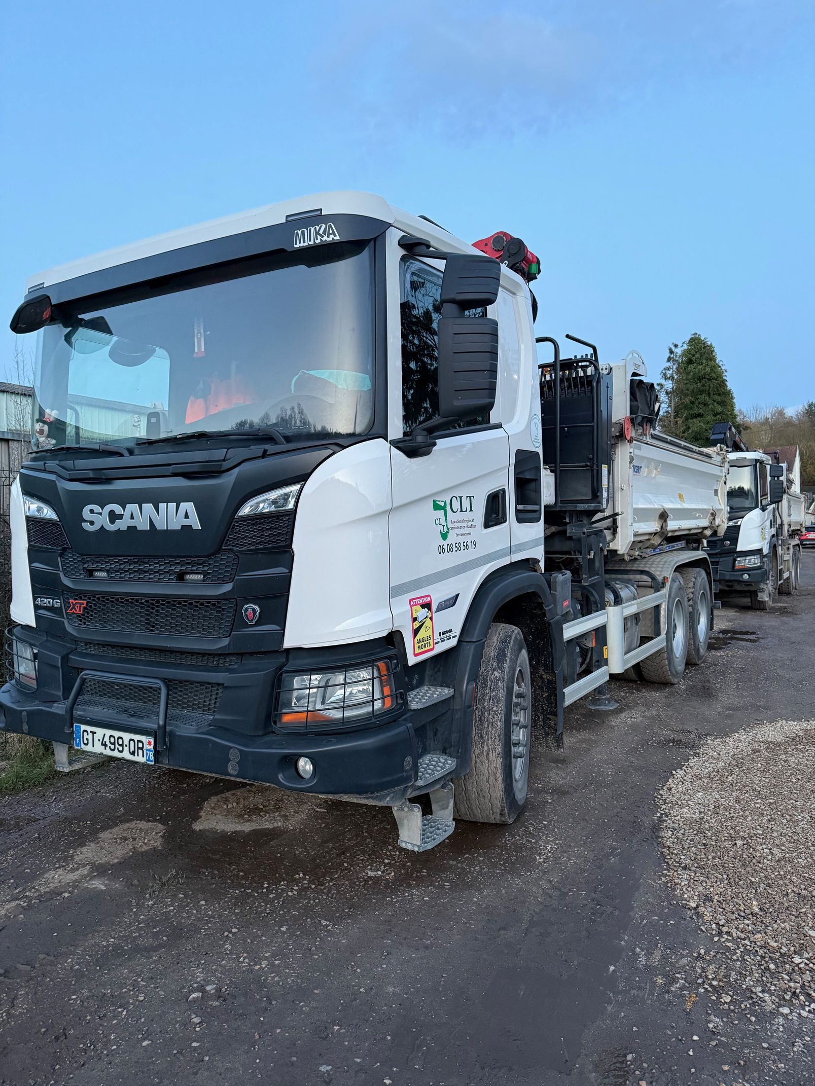 Un camion-benne Scania blanc, pneus couverts de boue, est stationné sur un sol gravillonné. Un autre camion est visible à l'arrière-plan.