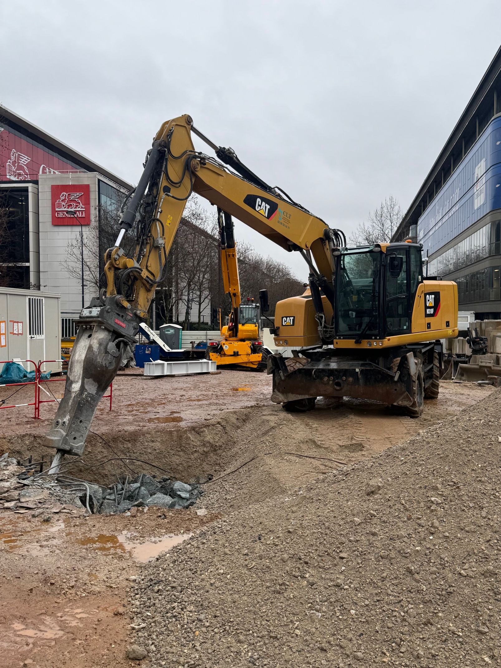 Une pelleteuse jaune casse du béton dans une zone de construction près de bâtiments par temps nuageux.