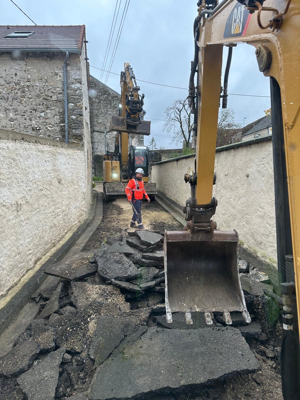 Chantier avec des excavatrices et un ouvrier qui arrachent l'asphalte d'une rue étroite.