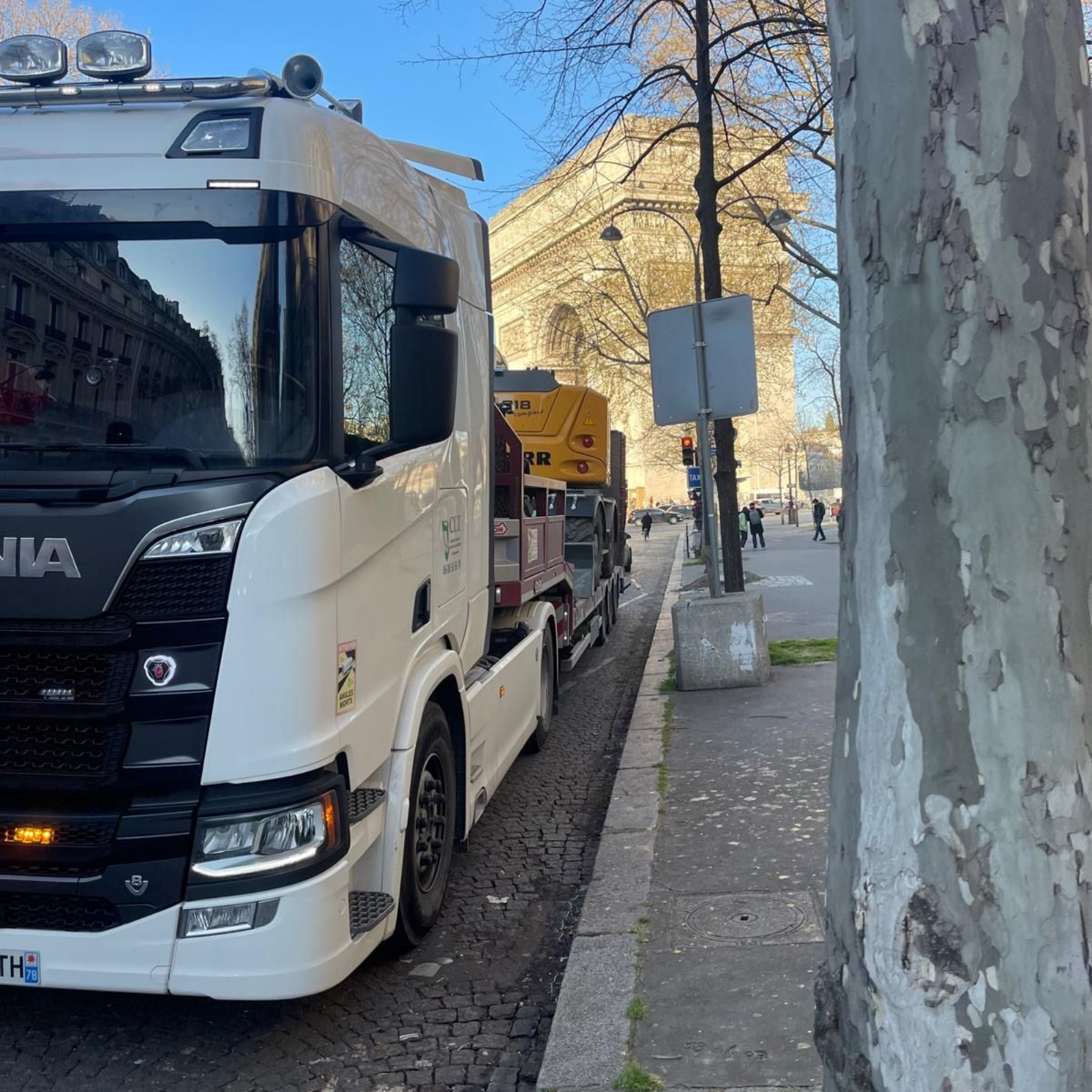 Camion Scania blanc avec équipement BTP sur une remorque, stationné dans une rue de la ville.