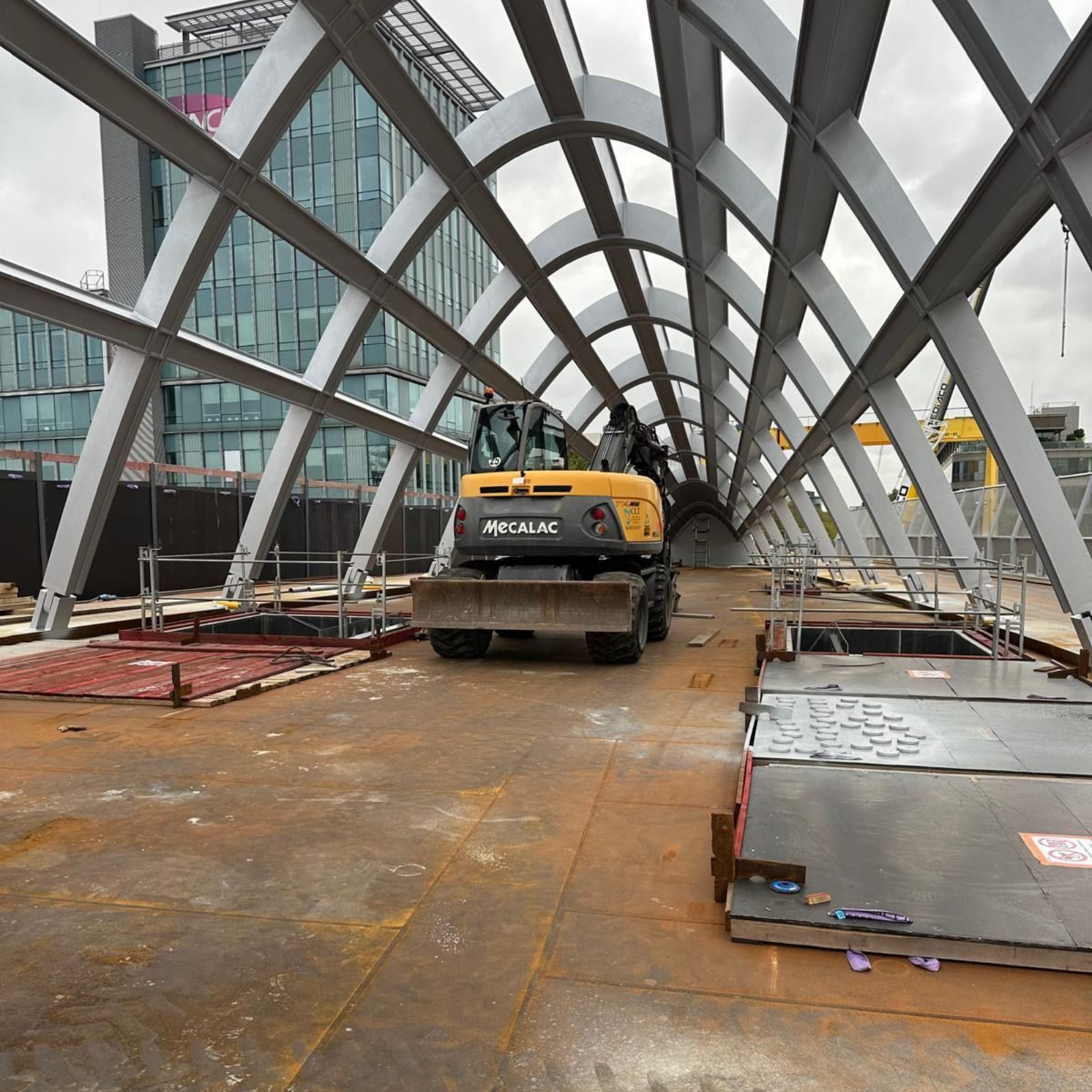 Chantier de construction avec une pelleteuse jaune sur un pont métallique en arc. Des poutres d'acier sont visibles ; un bâtiment se dresse à l'arrière-plan.