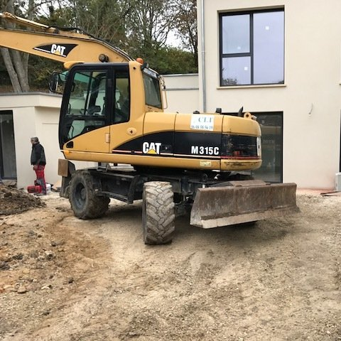 Une pelle mécanique CAT M315C jaune se trouve sur un chantier de construction, à proximité d'un bâtiment. Une personne se tient à proximité.